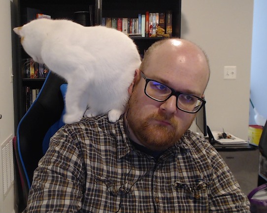 The author's calico cat sitting on his shoulder, while the author scowls with annoyance, despite internally feeling loved like he has never felt before